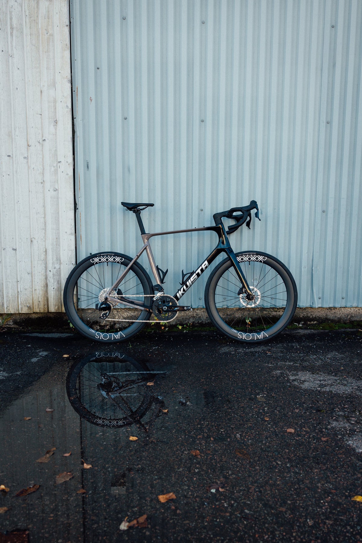 IKUISTA Spektri road bike leaning against a corrugated metal wall, reflected in a wet pavement puddle